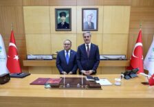 Görselde iki yönetici, belediye odasında duruyor, arka planda Türk bayrakları ve resmi portreler bulunuyor.